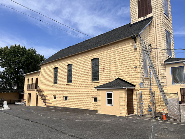Side view of a yellow stone church building with large windows, a tower, and a small entrance, set against a clear blue sky.