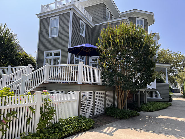 Exterior view of a stylish gray house with a white fence, featuring a shaded deck with a blue umbrella and lush greenery.