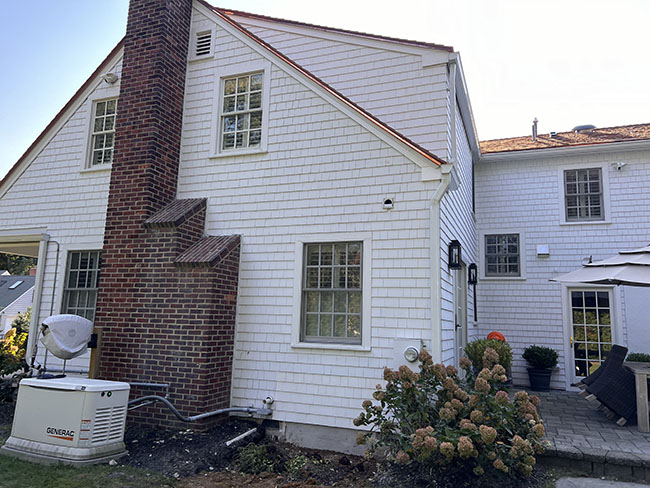 A white shingle-clad house with a brick chimney, an outdoor generator, and a patio area complete with potted plants and seating.