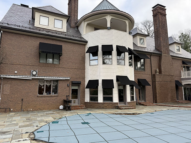 Exterior view of a large, modern home featuring a mix of brick and white siding, black awnings, and a pool cover on the stone patio.