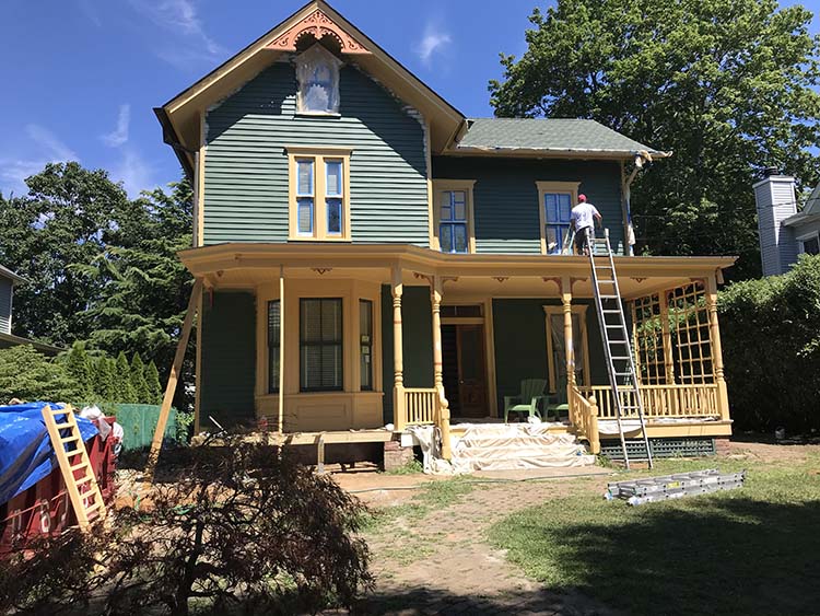A worker paints a Victorian-style house with green siding and yellow trim, using a ladder on a sunny day.