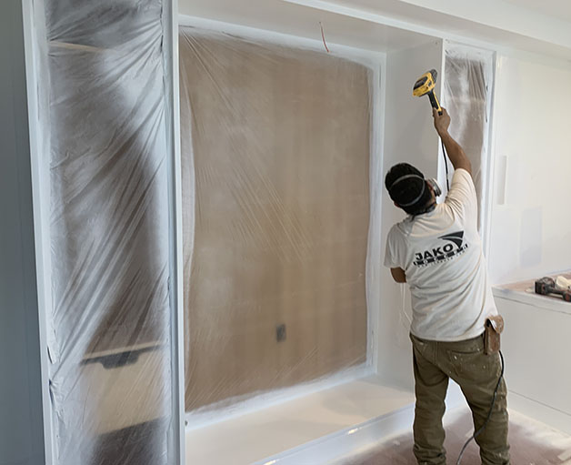 A worker in a white shirt sands a wall covered in plastic sheeting, preparing for painting in a bright, unfinished room.