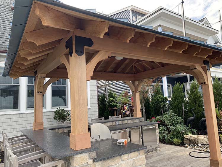 A wooden gazebo with a slate roof, featuring sturdy beams and a stone counter, surrounded by greenery in a residential backyard.