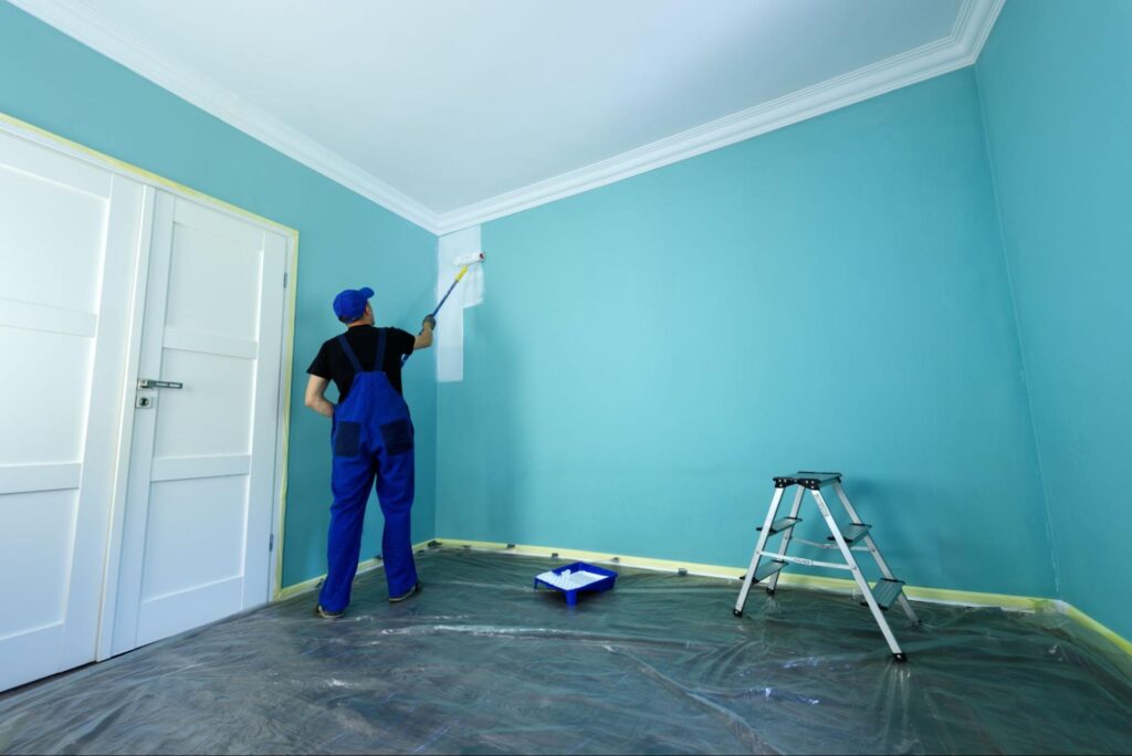 A male residential painter in overalls primes the Tiffany-blue wall for a new paint job; a ladder and paint tray are nearby.