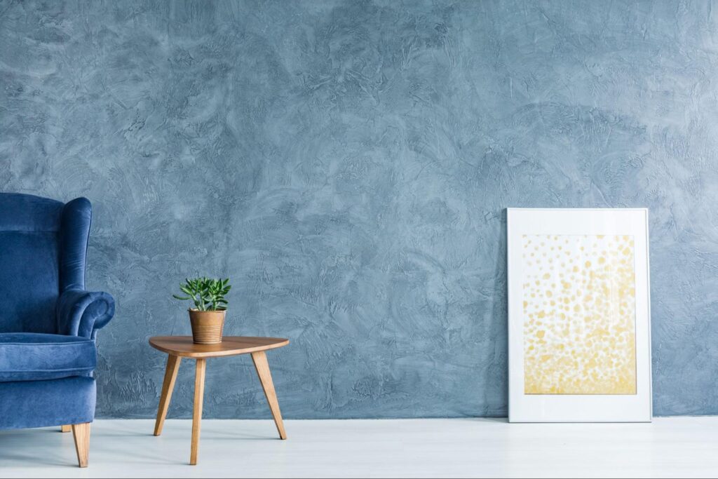 A minimalistic room featuring a blue couch, a round wooden table, and a white-framed artwork with gold designs against a textured blue wall.