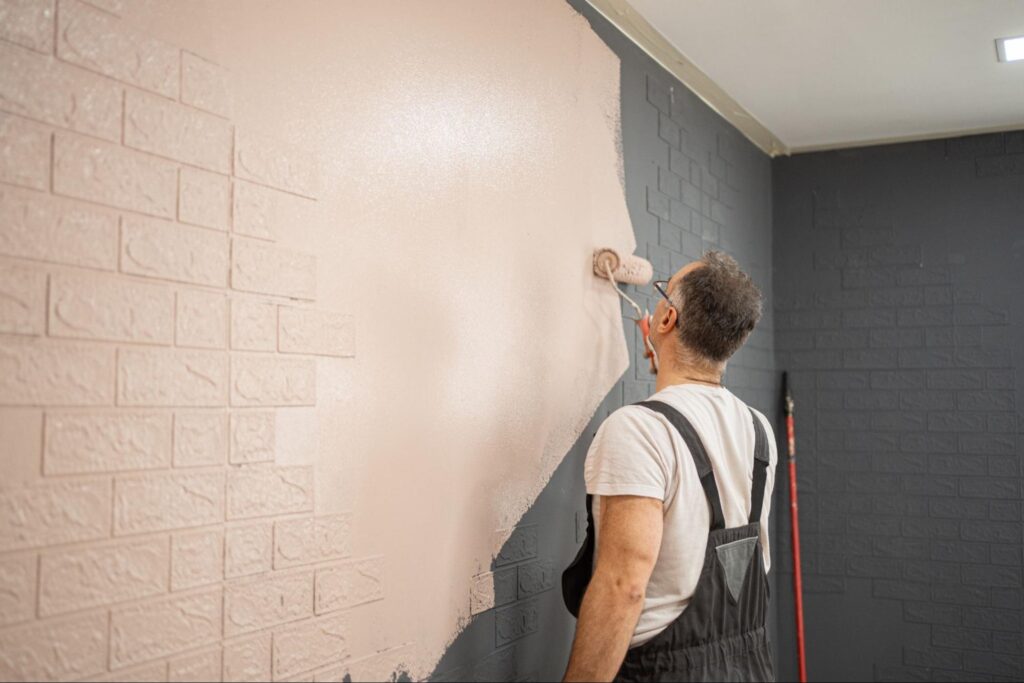 A residential painter uses a roller to apply peach paint to a gray brick wall.