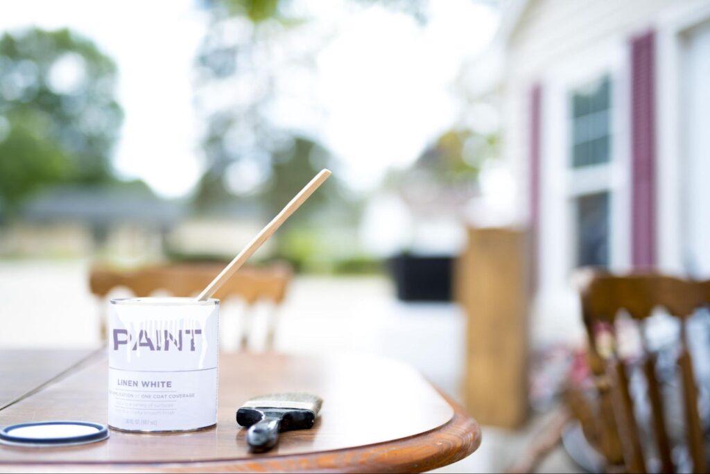 A can of paint and paintbrushes are on a table outside of a home.