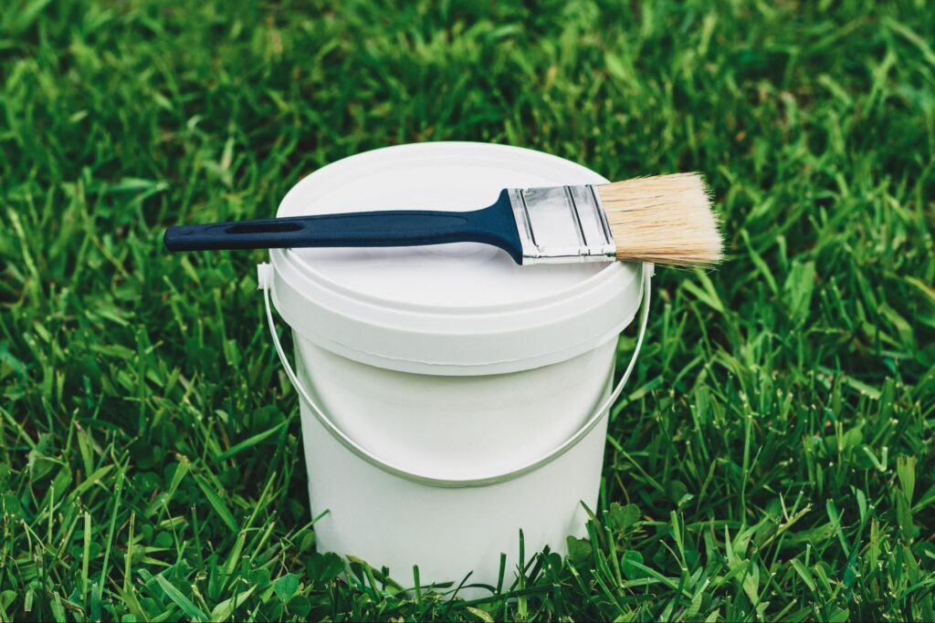 Paint brush and a bucket of paint on grass.