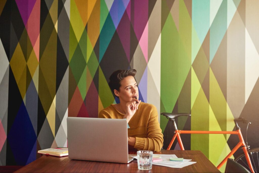 A woman at her desk, laptop open, framed by a bold geometric wall painted with vibrant, modern interior paint colors.