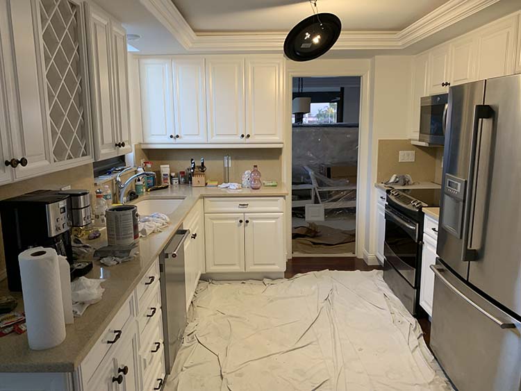 A cluttered kitchen with white cabinets, a stainless steel fridge, and a floor covered in a protective tarp, indicating renovation work.