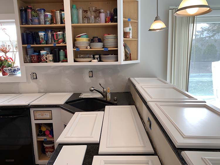 A kitchen undergoing renovation, featuring disassembled cabinet doors, cluttered shelves with dishes, and a sink area.
