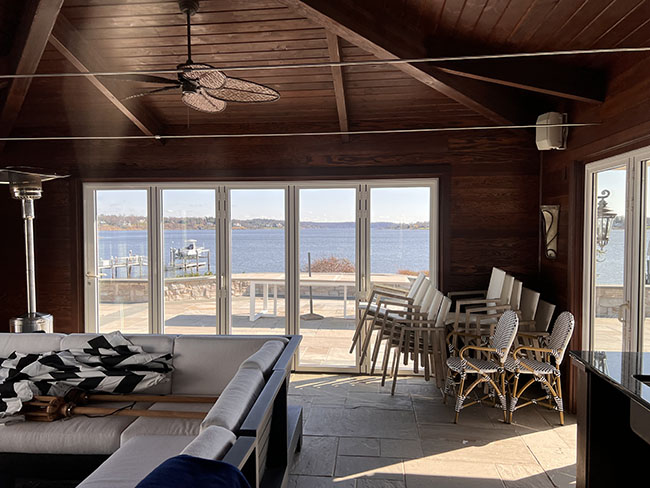 Cozy interior with a waterfront view, featuring white sofas, stacked chairs, and a fan under a wooden ceiling.