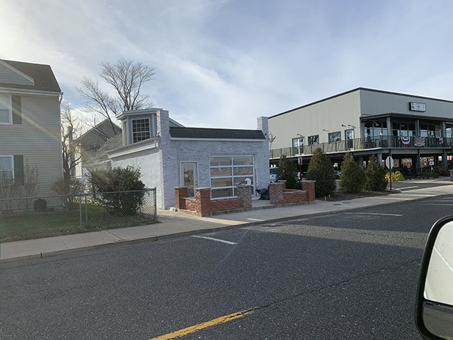 A small, white, brick building sits next to a larger commercial structure, set against a clear blue sky.