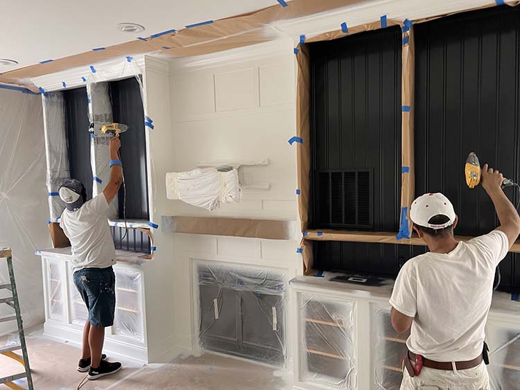 Two painters work on a room with partially covered walls, using tape and plastic sheeting, while one paints black paneling.