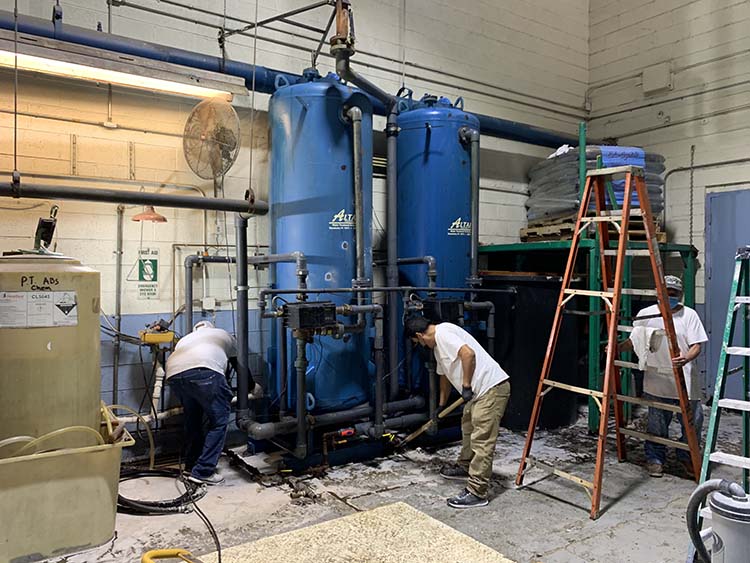 Three workers in white shirts maintain large blue tanks and pipes in a workshop, with a ladder and tools nearby.