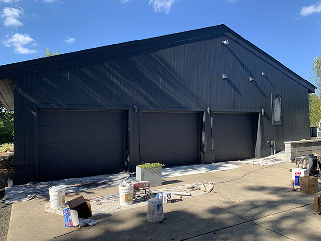 A newly painted black garage with three closed doors, surrounded by paint supplies on a sunny day.